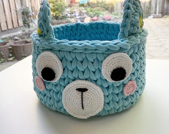 May include: A light blue crocheted storage basket shaped like a bear's face. The basket has white and black eyes, a white nose, and pink cheeks.