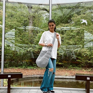 May include: A woman wearing a white t-shirt and blue jeans is carrying a gray crocheted tote bag over her shoulder. The bag has a long strap and is made of thick yarn.