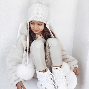 May include: A young girl wearing a white knitted hat with a pom-pom, a white faux fur jacket, white pants, and white snow boots.