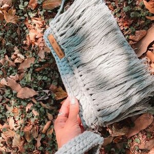 May include: A light blue crocheted bag with a fringe detail and a brown leather tag that says "Cool Like." The bag is held in a hand with a bracelet.
