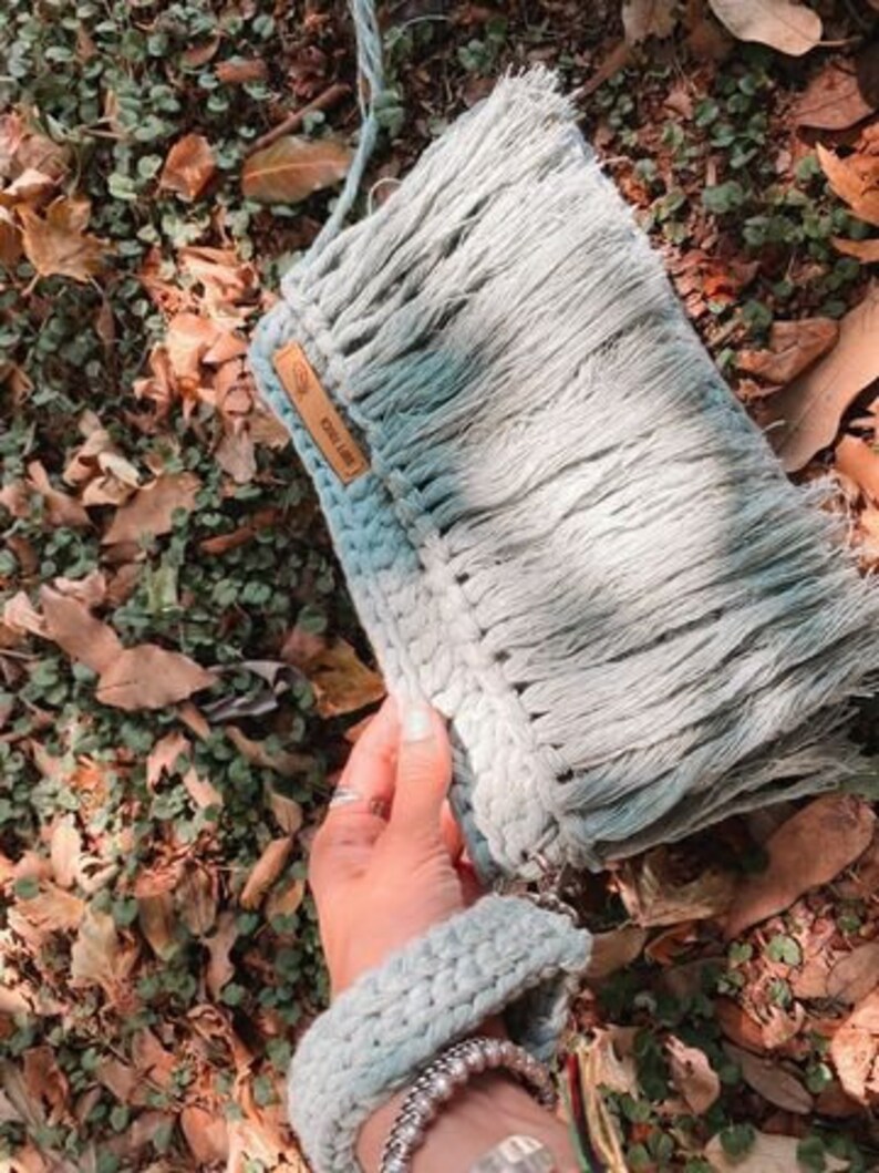 May include: A light blue crocheted bag with a fringe detail and a brown leather tag that says "Cool Like." The bag is held in a hand with a bracelet.