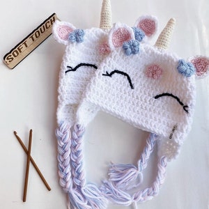 May include: Two white crocheted unicorn hats with pink and blue flower accents. The hats have long braids with pink, blue, and white yarn.