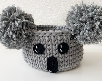 May include: Gray crocheted koala-shaped basket with black eyes and nose. The basket has two large pom-poms on the top.