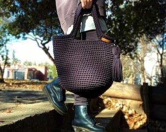 May include: A dark purple crocheted tote bag with a wooden handle and a long tassel. The bag is being carried by a person wearing black boots.