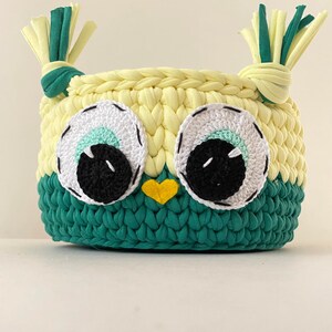 May include: A crocheted owl-shaped basket with green and yellow yarn. The owl has large white eyes with black pupils and a yellow beak.
