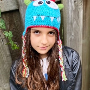 K&ouml;nnte beinhalten: Ein Kind tr&auml;gt eine blaue, geh&auml;kelte Monsterm&uuml;tze mit wei&szlig;en Z&auml;hnen, gr&uuml;nen H&ouml;rnern und roten und wei&szlig;en geflochtenen Garnb&auml;ndern. Die M&uuml;tze hat wei&szlig;e Augen und einen roten Rand.