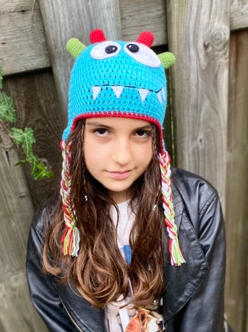 K&ouml;nnte beinhalten: Ein Kind tr&auml;gt eine blaue, geh&auml;kelte Monsterm&uuml;tze mit wei&szlig;en Z&auml;hnen, gr&uuml;nen H&ouml;rnern und roten und wei&szlig;en geflochtenen Garnb&auml;ndern. Die M&uuml;tze hat wei&szlig;e Augen und einen roten Rand.