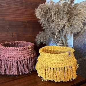 May include: Two crocheted baskets with fringe, one is brown and the other is yellow. The baskets are sitting on a wooden surface.