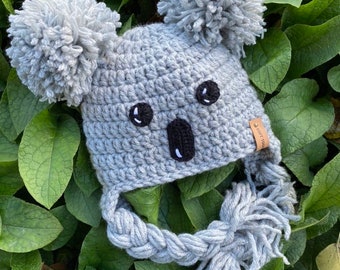 May include: Gray crocheted koala hat with black eyes and nose, pom poms on top, and a long braided tassel. The hat is lying on a bed of green leaves.