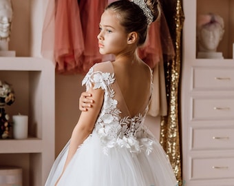 May include: A young girl wearing a white tulle dress with floral lace detailing and a low back. The dress has a full skirt and is perfect for a special occasion.