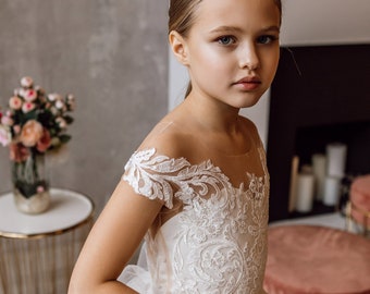 May include: A young girl wearing a white tulle dress with lace detailing. The dress has a sweetheart neckline and short sleeves. The girl is standing in front of a fireplace.