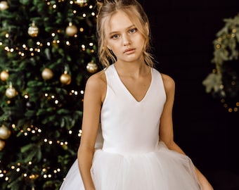 May include: A young girl wearing a white sleeveless dress with a tulle skirt. She is standing in front of a Christmas tree with gold ornaments and lights. The text "Boutique De Charme" is written in white on a black background.