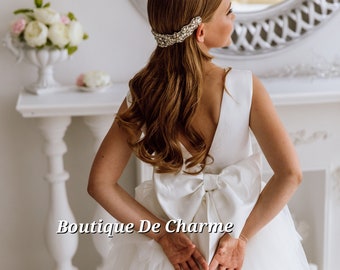 May include: A white bridal gown with a large bow in the back. The gown has a tulle skirt and a fitted bodice. The model is wearing a crystal hairpiece.