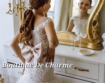 May include: A young girl in a white dress with a large pink bow on the back. The dress has lace detailing and a tulle skirt. The girl is standing in front of a vanity with a mirror and candles.