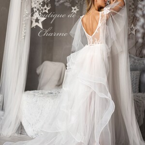 May include: A white tulle wedding dress with a lace bodice and a flowing skirt. The dress has a low back and a delicate lace design. The dress is being worn by a model in a studio setting.