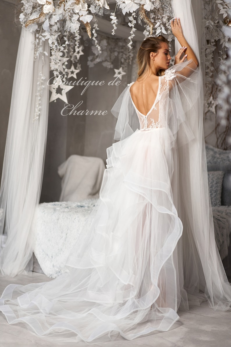 May include: A white tulle wedding dress with a lace bodice and a flowing skirt. The dress has a low back and a delicate lace design. The dress is being worn by a model in a studio setting.