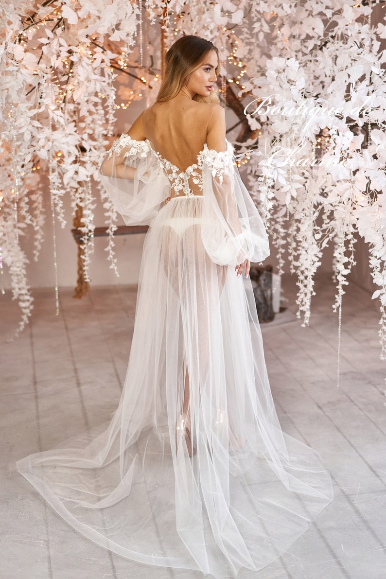 May include: A white sheer bridal robe with long sleeves and floral lace detailing. The robe has a flowing skirt and is worn over a white lingerie set.