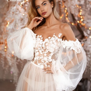 May include: A white sheer gown with a floral lace bodice and large, puffy sleeves. The gown is worn by a model with long blonde hair and a natural makeup look. The background is a blurred image of a white and gold Christmas tree.