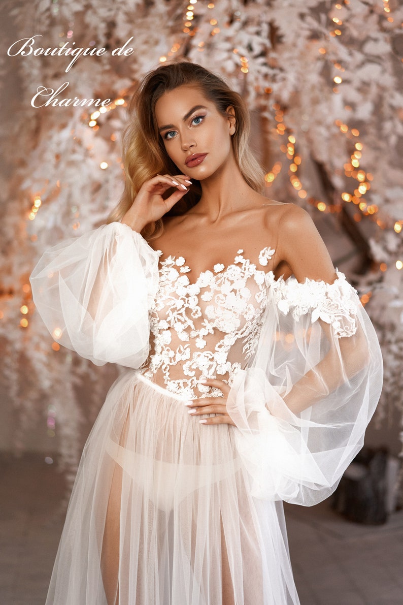 May include: A white sheer gown with a floral lace bodice and large, puffy sleeves. The gown is worn by a model with long blonde hair and a natural makeup look. The background is a blurred image of a white and gold Christmas tree.