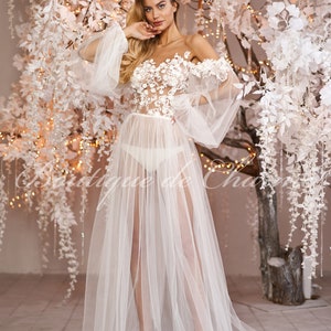 May include: A white sheer gown with a floral lace bodice and long, flowing sleeves. The gown is worn by a model standing in front of a white floral backdrop.