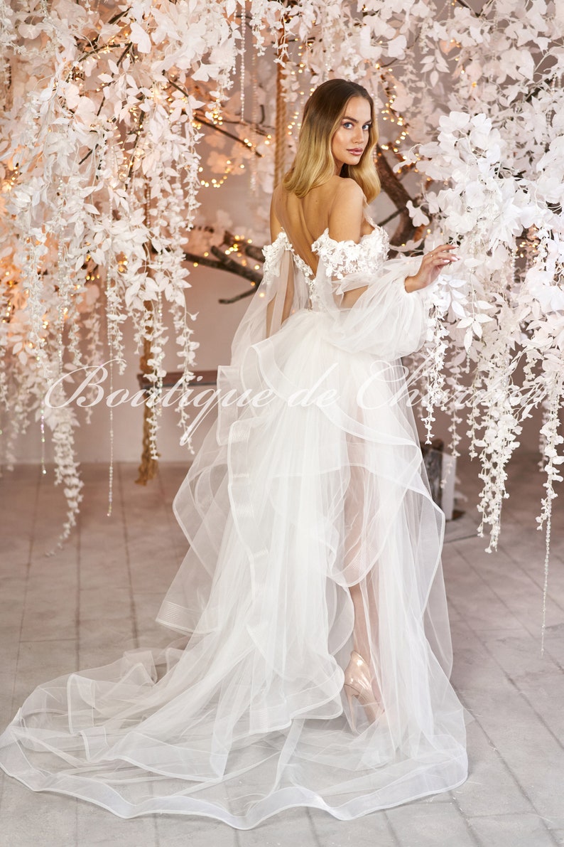 May include: A white wedding dress with a tulle skirt and off-the-shoulder lace bodice. The dress has long, puffy sleeves and a flowing train.