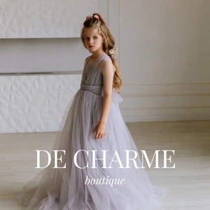 Puede incluir: Una niña pequeña con un vestido de tul gris claro y un accesorio floral para el cabello. El vestido tiene un corpiño ajustado y una falda fluida. El texto "DE CHARME boutique" está impreso debajo de la niña.