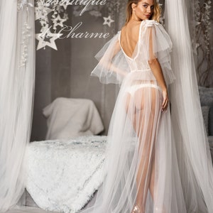 May include: A white sheer gown with lace detailing and a long train. The gown has a low back and is worn with gold heels. The model is standing in a bedroom with a white bed and a white canopy.
