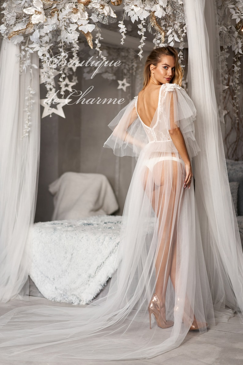 May include: A white sheer gown with lace detailing and a long train. The gown has a low back and is worn with gold heels. The model is standing in a bedroom with a white bed and a white canopy.