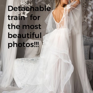 May include: A white wedding dress with a lace bodice and a flowing tulle skirt. The skirt has a detachable train, which can be removed for a more casual look. The dress is being worn by a model in a studio setting.