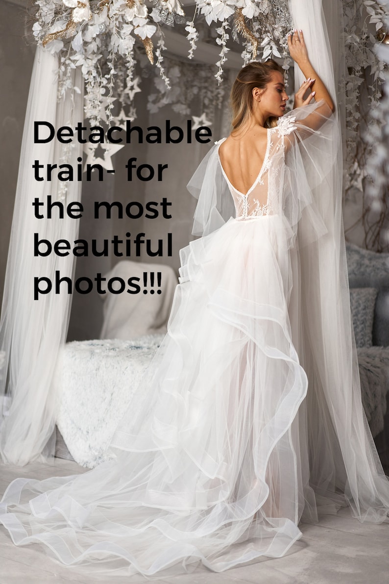 May include: A white wedding dress with a lace bodice and a flowing tulle skirt. The skirt has a detachable train, which can be removed for a more casual look. The dress is being worn by a model in a studio setting.