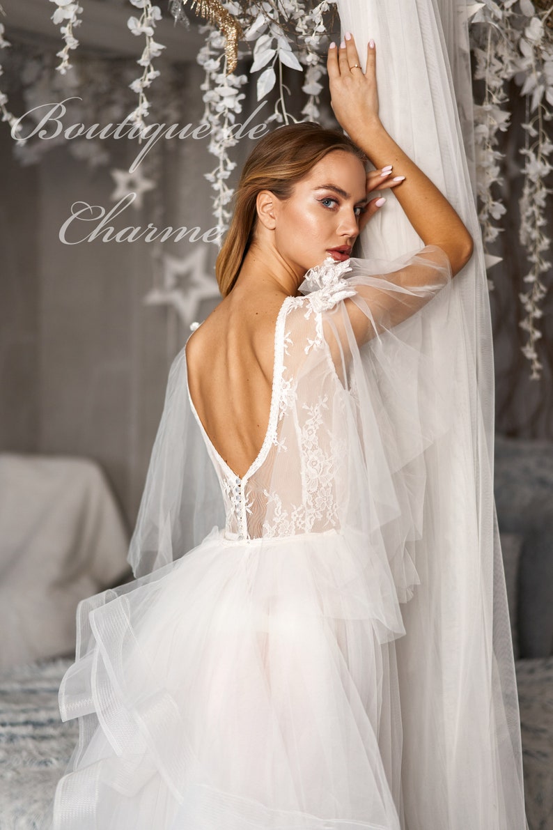 May include: A white wedding dress with a lace bodice and tulle skirt. The dress has a low back and a flowing skirt. The dress is being worn by a model in a studio setting.