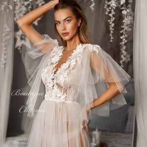 May include: A sheer white gown with delicate lace detailing and ruffled sleeves. The gown has a deep V-neckline and a flowing skirt.