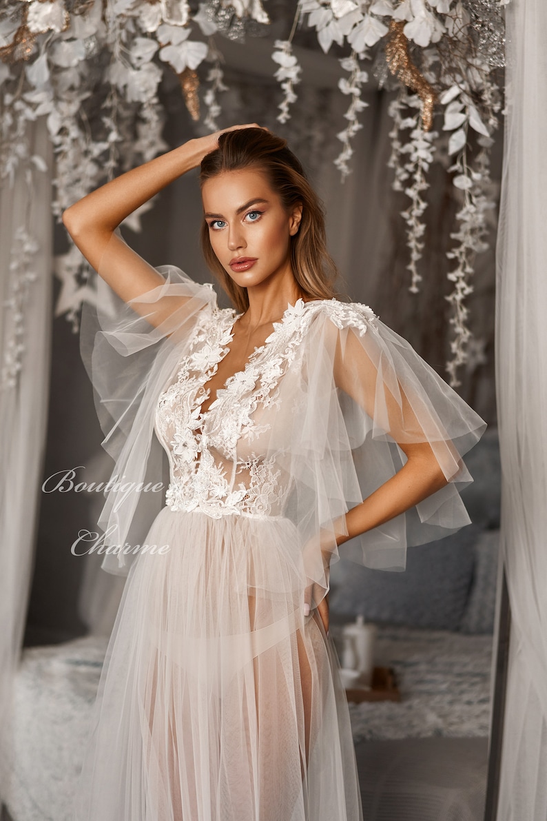 May include: A sheer white gown with delicate lace detailing and ruffled sleeves. The gown has a deep V-neckline and a flowing skirt.