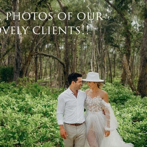 May include: A couple in a lush green forest setting. The woman is wearing a white off-the-shoulder dress with lace detailing and a white hat. The man is wearing a white button-down shirt and khaki pants. They are looking at each other and smiling.