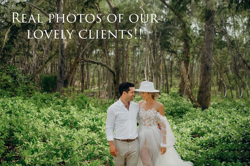 May include: A couple in a lush green forest setting. The woman is wearing a white off-the-shoulder dress with lace detailing and a white hat. The man is wearing a white button-down shirt and khaki pants. They are looking at each other and smiling.