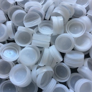 May include: A close-up shot of a pile of white plastic bottle caps. The caps have a circular design with a ridged edge and a flat top. Some caps are stacked, while others are scattered.