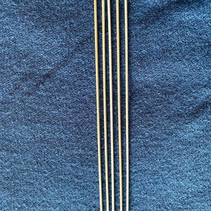 May include: Five thin, light brown wooden skewers arranged vertically against a textured, dark blue background. The skewers are evenly spaced and appear to be new.