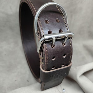 May include: A dark brown leather dog collar with a silver metal buckle and D-ring. The collar features visible stitching and multiple adjustment holes. The leather appears thick and durable, suggesting a high-quality product for pet owners.