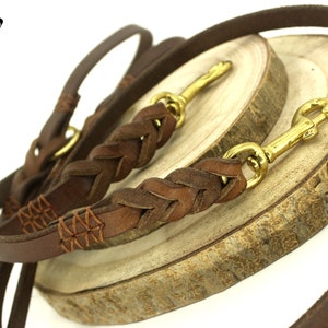 Pode incluir: Duas trelas de couro marrom para cães com detalhes trançados e costurados. As trelas têm fechos de metal dourado e são exibidas em uma superfície de madeira. "Heavy Duty Collars" está escrito em preto sobre fundo branco.