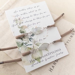 May include: Two matching friendship bracelets with a light blue oval gemstone charm. The bracelets are made of brown cord and have silver clasps. The bracelets are displayed on a white card with a floral design and the text "No matter where we go. No matter what we do. You'll always be there for me And I'll always be there for you One for you One for me HANDMADESTYLINGS Handmade Gemstone Jewelry HAND TYLINGS".