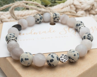 May include: A stretch bracelet featuring alternating matte white beads and speckled dalmatian jasper beads, accented with a silver-toned metal paw print charm and two silver-toned spacer rings, displayed on a white card with the text Handmade Stylings.