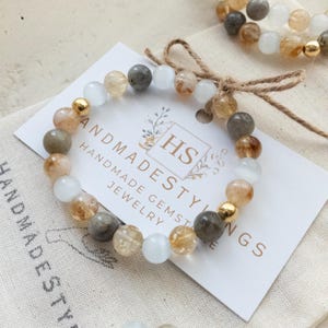 May include: A bracelet made with white, gray, and gold beads. The bracelet is on a white card with the text "Handmade Gemstone Jewelry" and "Handmadesythings" on it.
