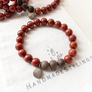 May include: A red gemstone bracelet with a spiral charm. The bracelet is made of round beads and is on a white fabric background. The text "HANDMADESTYNGS" is visible on the fabric.