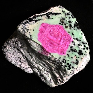 May include: A raw mineral specimen featuring a vibrant, hexagonal magenta crystal embedded within a matrix of pale mint green stone. The surface displays contrasting black speckled inclusions and dark, layered grey mineral formations along the side.