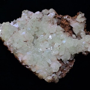 May include: A cluster of pale green crystals with a crystalline structure, set against a backdrop of brown and tan mineral formations. The crystals have a translucent quality, and the overall composition is set against a black background.