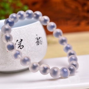 Puede incluir: Un brazalete de ágata blanca con inclusiones grises y azules. El brazalete está hecho de cuentas redondas y está enhebrado en un cordón elástico transparente.