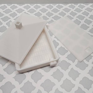May include: White plastic container with a lid. The container has a grid pattern on the bottom and a small square protrusion on the lid.