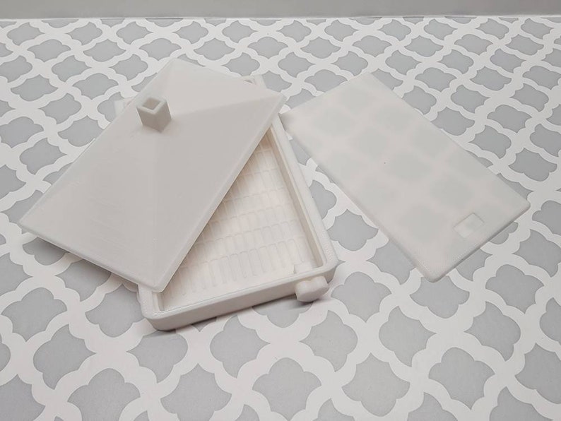 May include: White plastic container with a lid. The container has a grid pattern on the bottom and a small square protrusion on the lid.