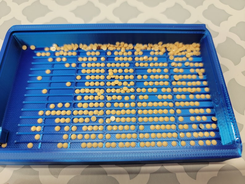 Puede incluir: Una bandeja de pl&aacute;stico azul con filas de peque&ntilde;as cuentas redondas de color beige. La bandeja est&aacute; llena de cuentas, y algunas est&aacute;n dispersas alrededor de los bordes.