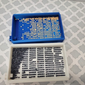 Puede incluir: Dos bandejas de pl&aacute;stico para organizar los taladros de pintura de diamantes. La bandeja superior es azul y tiene peque&ntilde;os taladros redondos de color beige. La bandeja inferior es blanca y tiene filas de peque&ntilde;os taladros cuadrados negros.